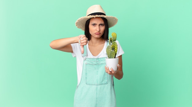 Bonita mujer campesina que se siente cruzada, mostrando los pulgares hacia abajo y sosteniendo un cactus