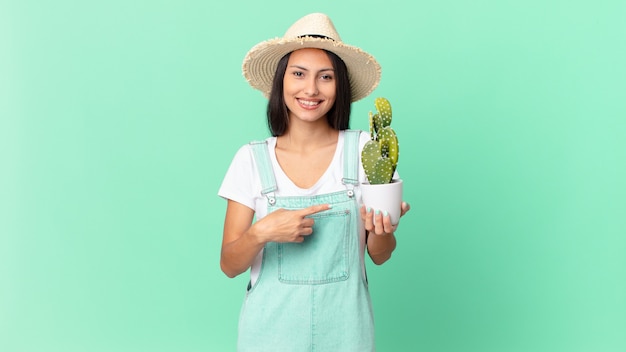 Bonita mujer campesina sonriendo alegremente, sintiéndose feliz y apuntando hacia un lado y sosteniendo un cactus