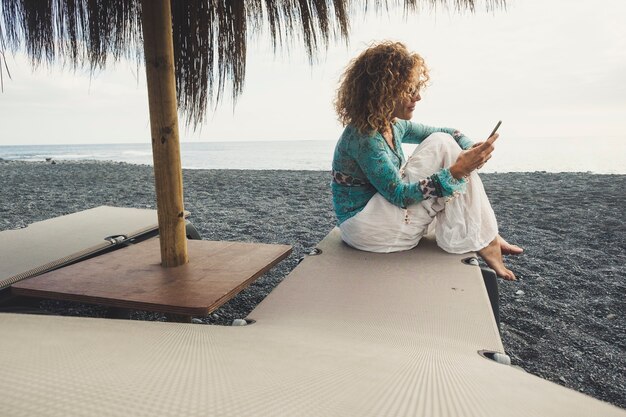 Bonita mujer caucásica de mediana edad en la playa, siéntese en los asientos con el océano y la arena a su alrededor, revise el teléfono para ver Internet y los correos electrónicos y trabaje fuera de la oficina, manténgase conectado