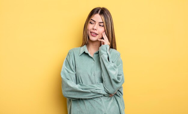 Bonita mujer caucásica sonriendo con una expresión feliz y segura con la mano en la barbilla, preguntándose y mirando hacia un lado