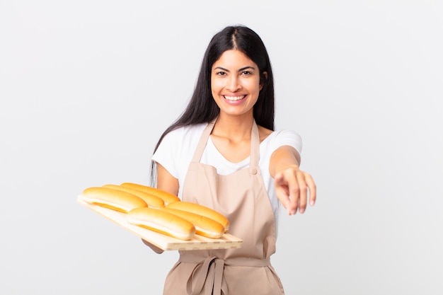 Bonita mujer chef hispana apuntando a la cámara eligiéndote y sosteniendo una bandeja con bollos de pan