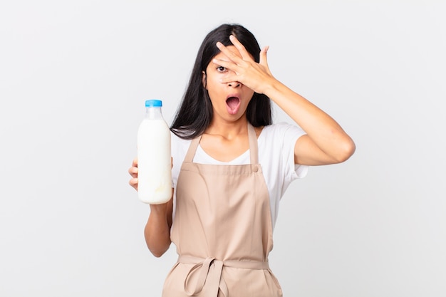 Bonita mujer chef hispana que parece sorprendida, asustada o aterrorizada, cubriéndose la cara con la mano y sosteniendo una botella de leche