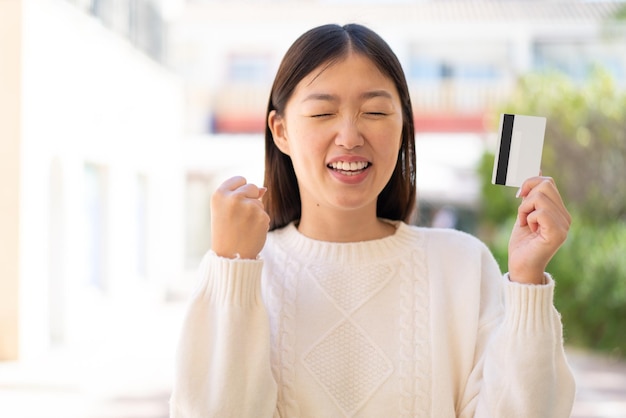 Bonita mujer china al aire libre sosteniendo una tarjeta de crédito y celebrando una victoria