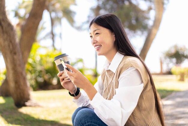 Bonita mujer china al aire libre usando un teléfono móvil y sosteniendo un café para llevar