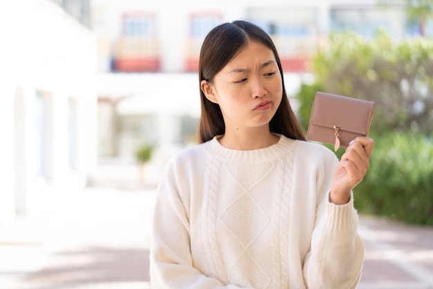 Bonita mujer china sosteniendo una billetera al aire libre con expresión triste