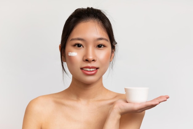 Bonita mujer coreana con una piel perfecta aplicando crema hidratante en la cara y sosteniendo un frasco con