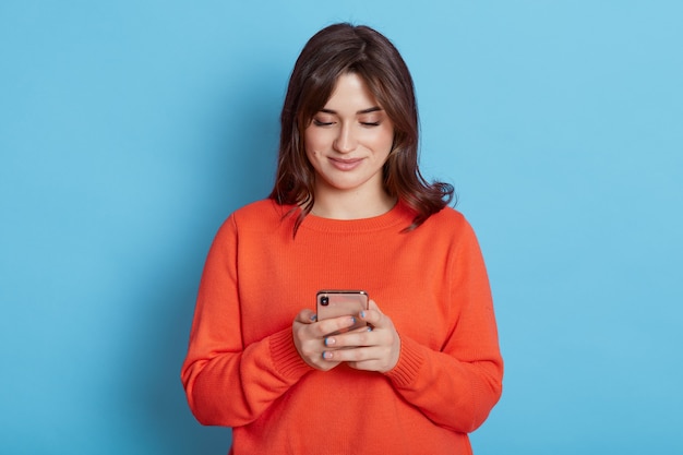 Bonita mujer europea con cabello oscuro, mira videos en un teléfono inteligente aislado en una pared azul, envía mensajes de texto, usa una aplicación especial en un dispositivo electrónico, sonríe suavemente, usa un jersey naranja casual.