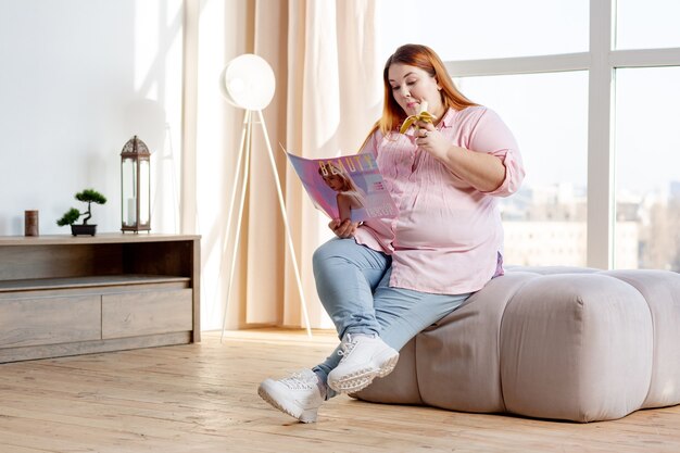 Bonita mujer hambrienta sosteniendo un plátano mientras lee una revista en casa