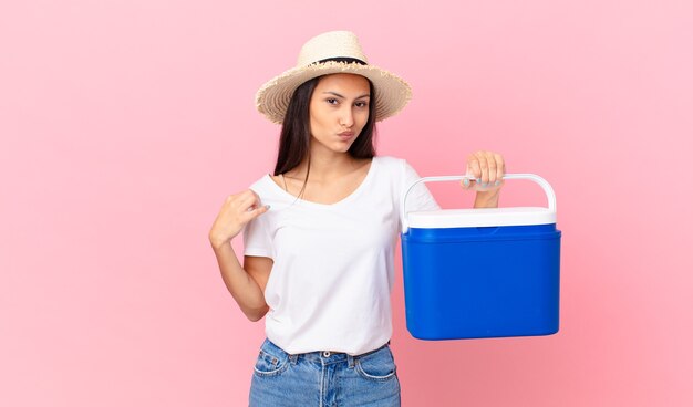 Bonita mujer hispana de aspecto arrogante, exitoso, positivo y orgulloso y sosteniendo un refrigerador portátil