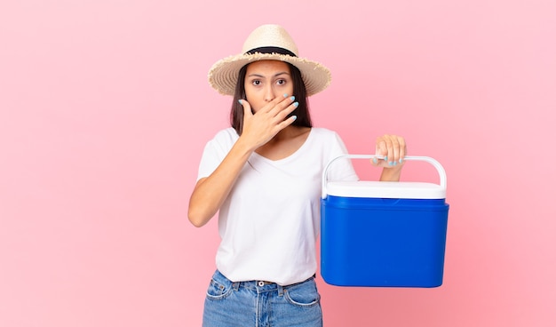 Bonita mujer hispana cubriendo la boca con las manos con un sorprendido y sosteniendo un refrigerador portátil