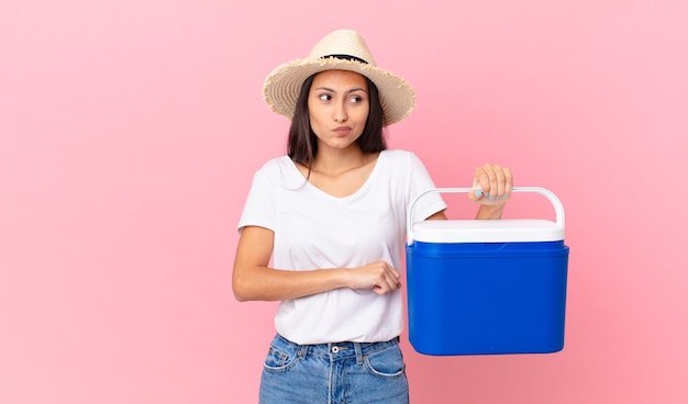 Bonita mujer hispana encogiéndose de hombros, sintiéndose confundida e insegura y sosteniendo un refrigerador portátil