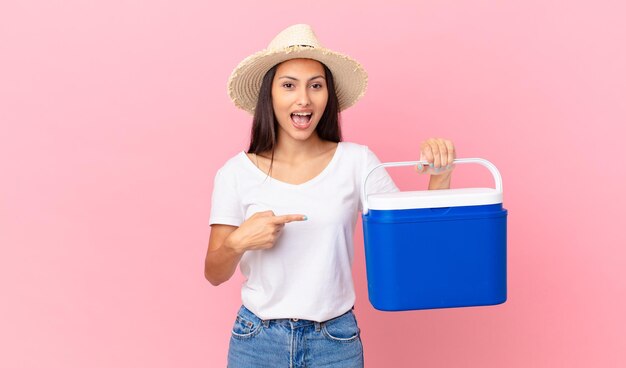 Bonita mujer hispana mirando emocionada y sorprendida apuntando hacia un lado y sosteniendo un refrigerador portátil