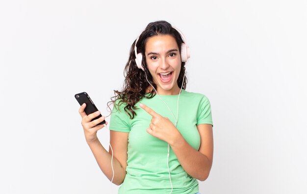 Bonita mujer hispana mirando emocionado y sorprendido apuntando hacia el lado con auriculares y un teléfono inteligente