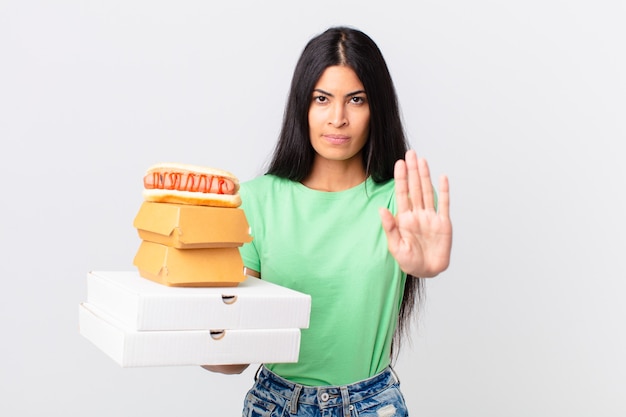 Bonita mujer hispana mirando seriamente mostrando la palma abierta haciendo gesto de parada y sosteniendo cajas de comida rápida para llevar