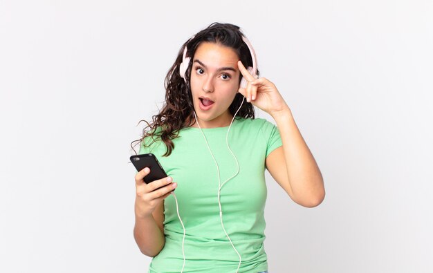 Bonita mujer hispana mirando sorprendida, dándose cuenta de un nuevo pensamiento, idea o concepto con auriculares y un teléfono inteligente