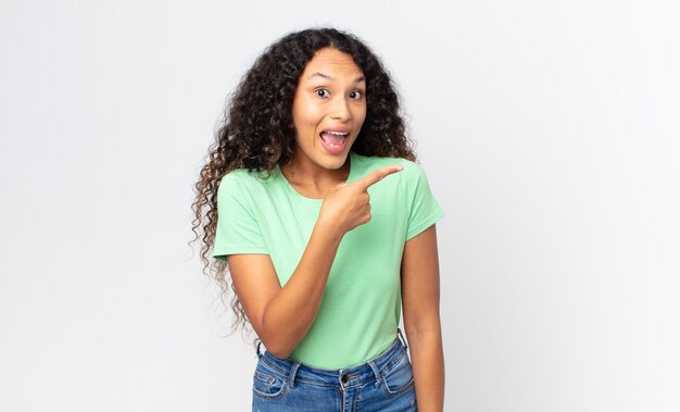 Bonita mujer hispana que parece emocionada y sorprendida apuntando hacia un lado y hacia arriba para copiar el espacio