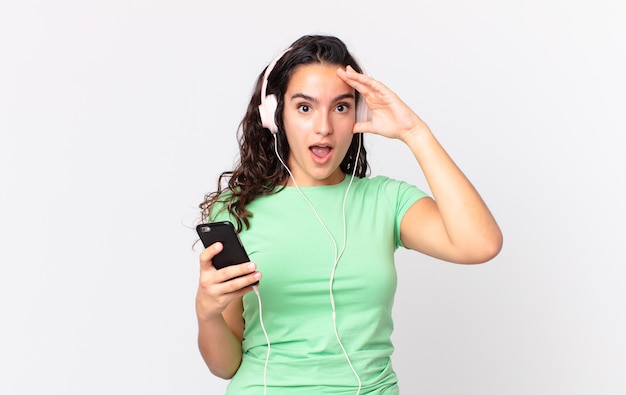 Bonita mujer hispana que parece feliz, asombrada y sorprendida con auriculares y un teléfono inteligente