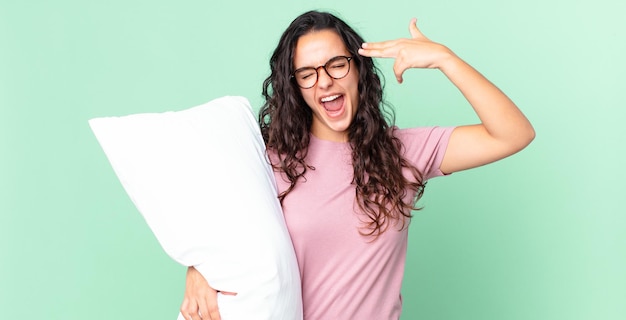 Foto bonita mujer hispana que parece infeliz y estresada, gesto de suicidio haciendo un signo de pistola y vistiendo pijama con una almohada