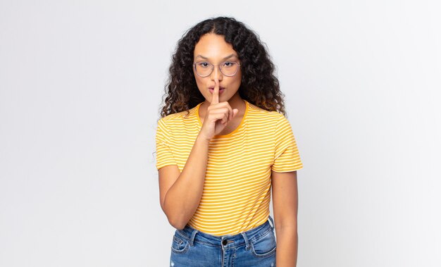 Bonita mujer hispana que parece seria y cruzada con el dedo presionado a los labios exigiendo silencio o silencio, guardando un secreto