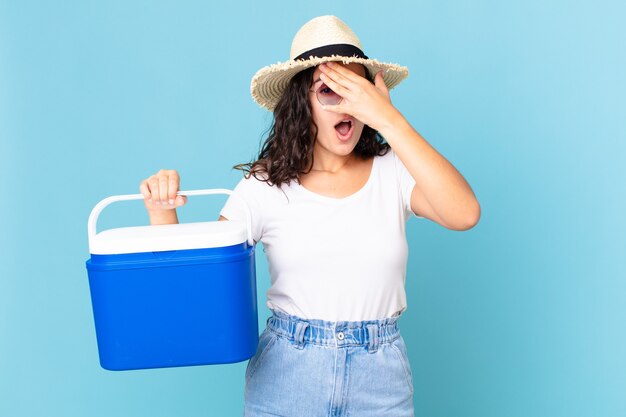 Foto bonita mujer hispana que parece sorprendida, asustada o aterrorizada, cubriendo la cara con la mano sosteniendo un refrigerador portátil