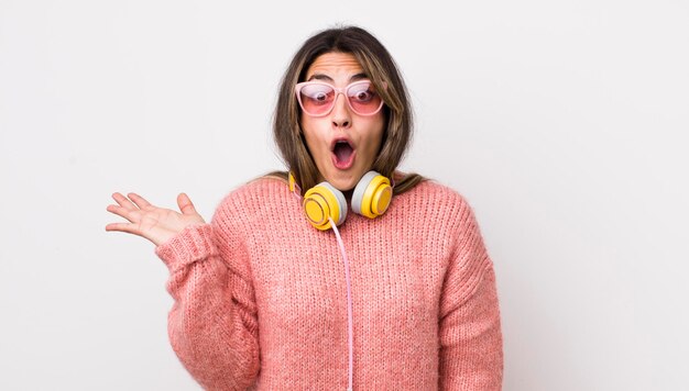 Bonita mujer hispana que parece sorprendida y conmocionada con la mandíbula caída sosteniendo un concepto de auriculares de objeto