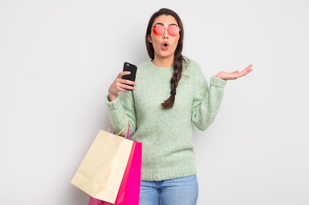 Bonita mujer hispana que parece sorprendida y conmocionada con la mandíbula caída sosteniendo un objeto bolsas de compras y concepto de cliente