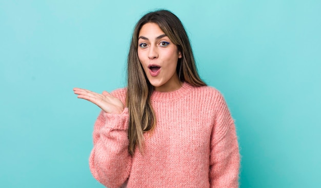 Bonita mujer hispana que parece sorprendida y conmocionada con la mandíbula caída sosteniendo un objeto con la mano abierta en el costado