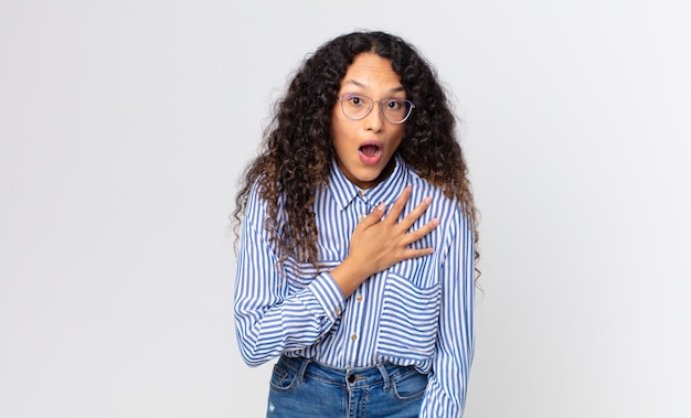 Bonita mujer hispana que se siente conmocionada y sorprendida, sonriendo, tomando la mano al corazón, feliz de ser la indicada o mostrando gratitud