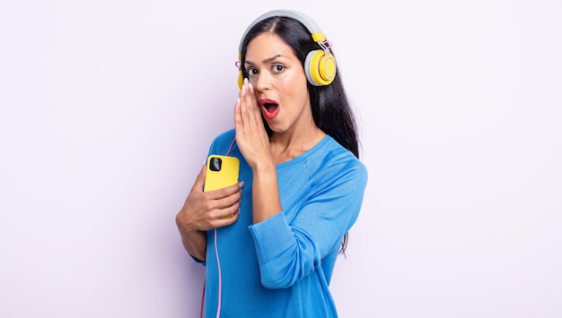 Bonita mujer hispana que se siente feliz, dando un gran grito con las manos al lado de la boca. concepto de teléfono inteligente y auriculares