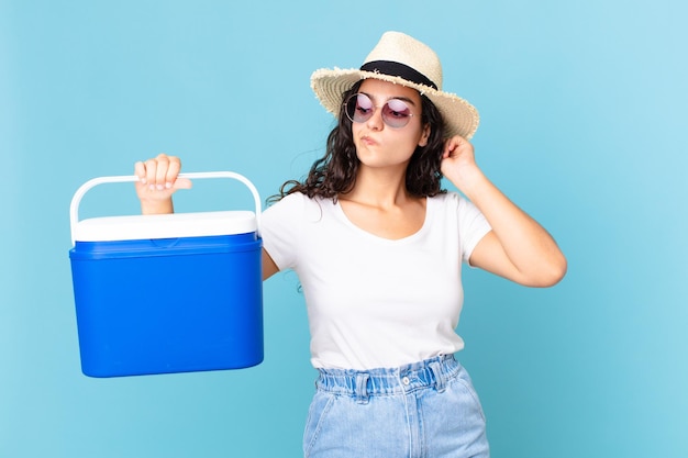 Bonita mujer hispana que se siente perpleja y confundida, rascándose la cabeza sosteniendo un refrigerador portátil