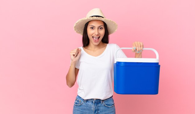 Bonita mujer hispana sintiéndose conmocionada, riendo y celebrando el éxito y sosteniendo un refrigerador portátil