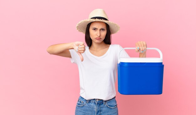 Bonita mujer hispana sintiéndose cruzada, mostrando los pulgares hacia abajo y sosteniendo un refrigerador portátil