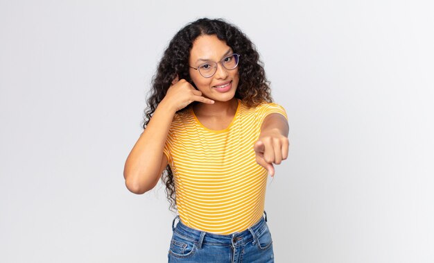 Bonita mujer hispana sonriendo alegremente y apuntando a la cámara mientras hace una llamada, gesto más tarde, hablando por teléfono