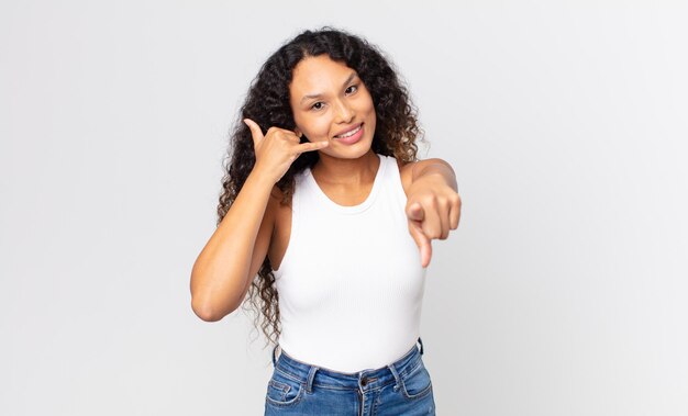 Bonita mujer hispana sonriendo alegremente y apuntando a la cámara mientras hace una llamada, gesto más tarde, hablando por teléfono