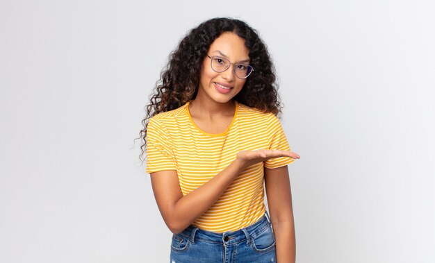 Bonita mujer hispana sonriendo alegremente, sintiéndose feliz y mostrando un concepto en el espacio de la copia con la palma de la mano