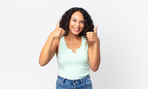 Bonita mujer hispana sonriendo con alegría y luciendo feliz, sintiéndose despreocupada y positiva con ambos pulgares hacia arriba