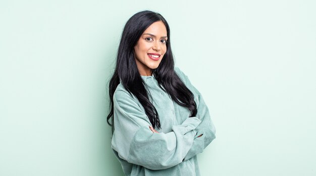 Bonita mujer hispana sonriendo a la cámara con los brazos cruzados y una expresión feliz, confiada y satisfecha, vista lateral