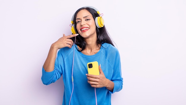 Bonita mujer hispana sonriendo con confianza apuntando a su propia sonrisa amplia. concepto de teléfono inteligente y auriculares