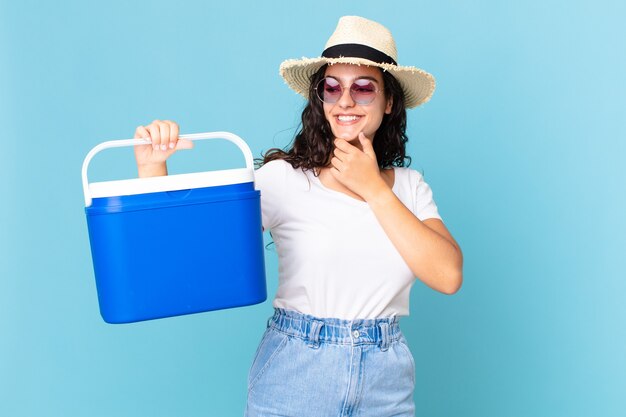 Bonita mujer hispana sonriendo con una expresión feliz y segura con la mano en la barbilla sosteniendo un refrigerador portátil