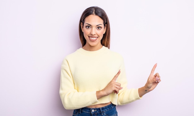 Bonita mujer hispana sonriendo felizmente y apuntando hacia un lado y hacia arriba con ambas manos mostrando el objeto en el espacio de la copia