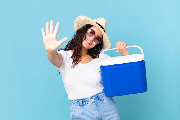 Foto bonita mujer hispana sonriendo y mirando amigable, mostrando el número cinco sosteniendo un refrigerador portátil