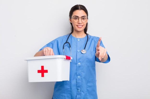 Bonita mujer hispana sonriendo con orgullo y confianza haciendo el número uno. enfermera y botiquín de primeros auxilios