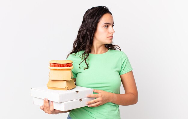 Bonita mujer hispana en la vista de perfil pensando, imaginando o soñando despierta y sosteniendo cajas de comida rápida para llevar