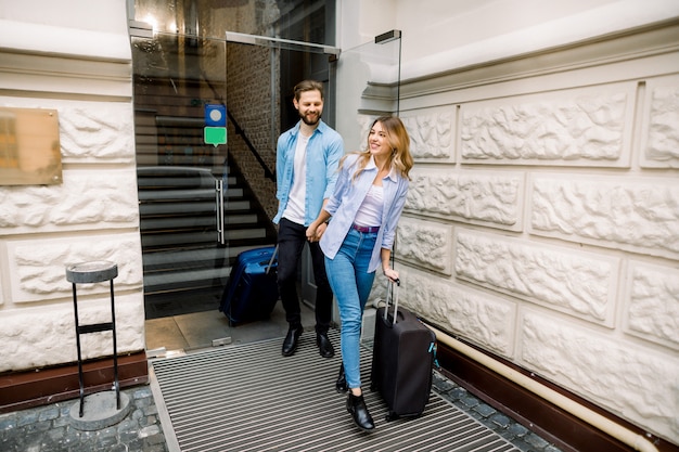 Bonita mujer y hombre guapo cogidos de la mano están saliendo de un hotel con sus maletas