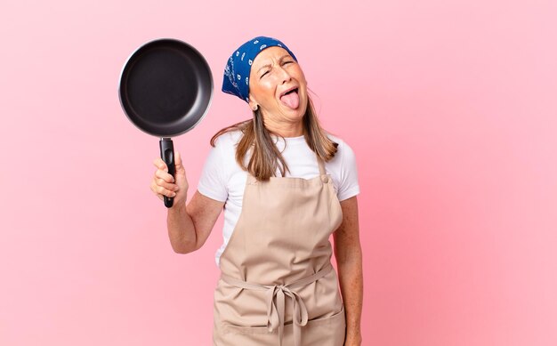 Bonita mujer de mediana edad con actitud alegre y rebelde, bromeando y sacando la lengua y sosteniendo una sartén. concepto de chef