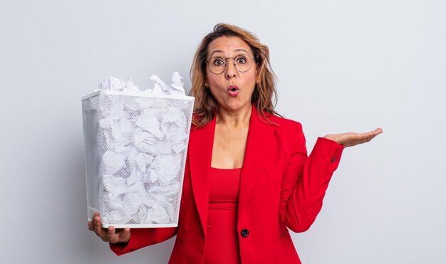 Bonita mujer de mediana edad que parece sorprendida y consternada, con la mandíbula caída sosteniendo un objeto. concepto de papelera de bolas de papel