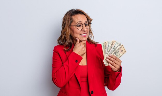 Bonita mujer de mediana edad sonriendo con una expresión feliz y segura con la mano en la barbilla. concepto de billetes de dólar