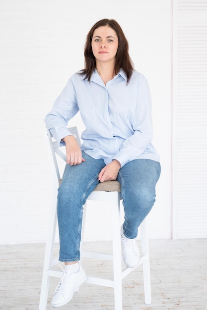 Bonita mujer modelo plus size en camisa y jeans posando en estudio en silla, fondo blanco con espacio de copia.