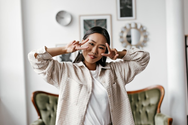 Bonita mujer morena con camiseta blanca y cárdigan beige sonríe sinceramente y muestra vsigns Atractiva dama con elegante atuendo posa en la sala de estar con sofá verde y cuadros en la pared blanca