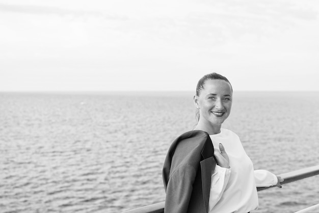 Bonita mujer de negocios o linda chica feliz con cara sonriente en blusa blanca chaqueta gris tiene manicura roja en la cubierta del barco viendo el agua del océano en el fondo del cielo nublado viajes y turismo